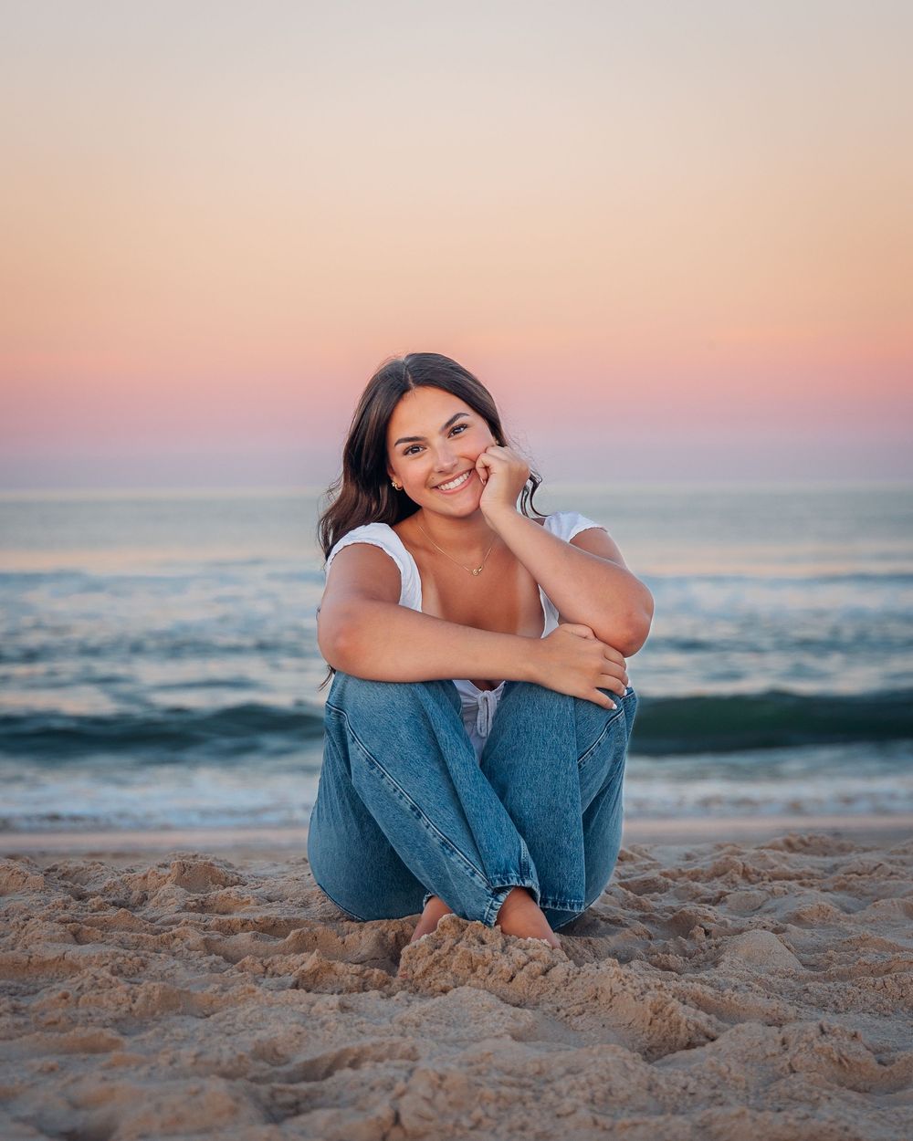 Young woman sitting on sandy beach at sunset, smiling in white top and jeans with ocean waves behind her.