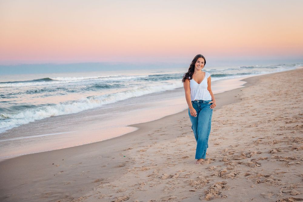 Woman in white top and jeans walking barefoot on sandy beach at sunset with gentle ocean waves.