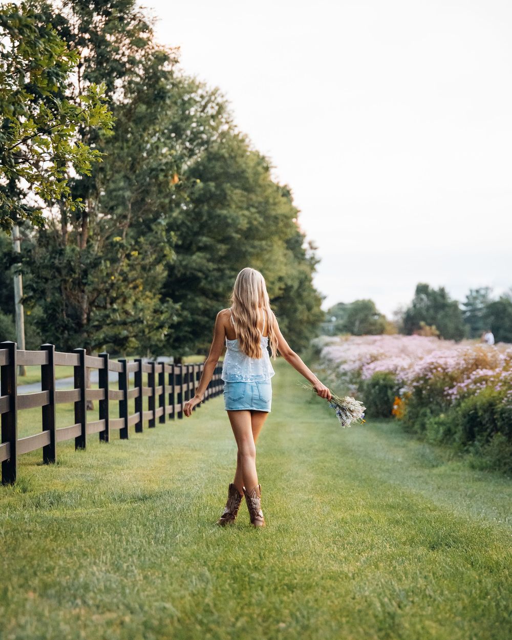 A young woman in denim skirt and cowboy boots walks along a grassy path beside a wooden fence.