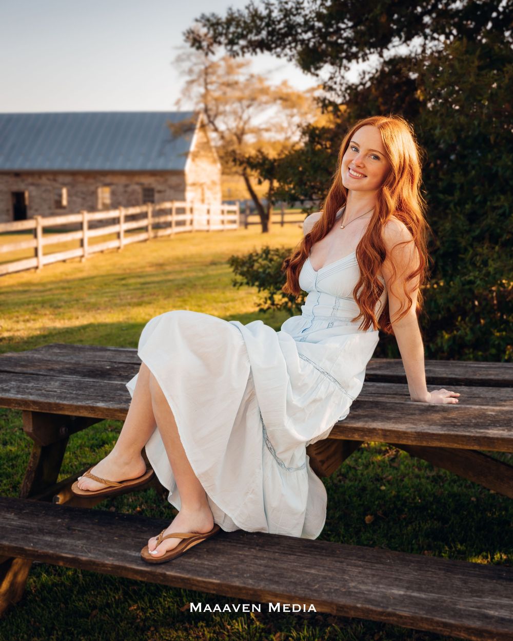 Young woman with long red hair in a white dress sitting on a wooden bench at a farm during golden hour.