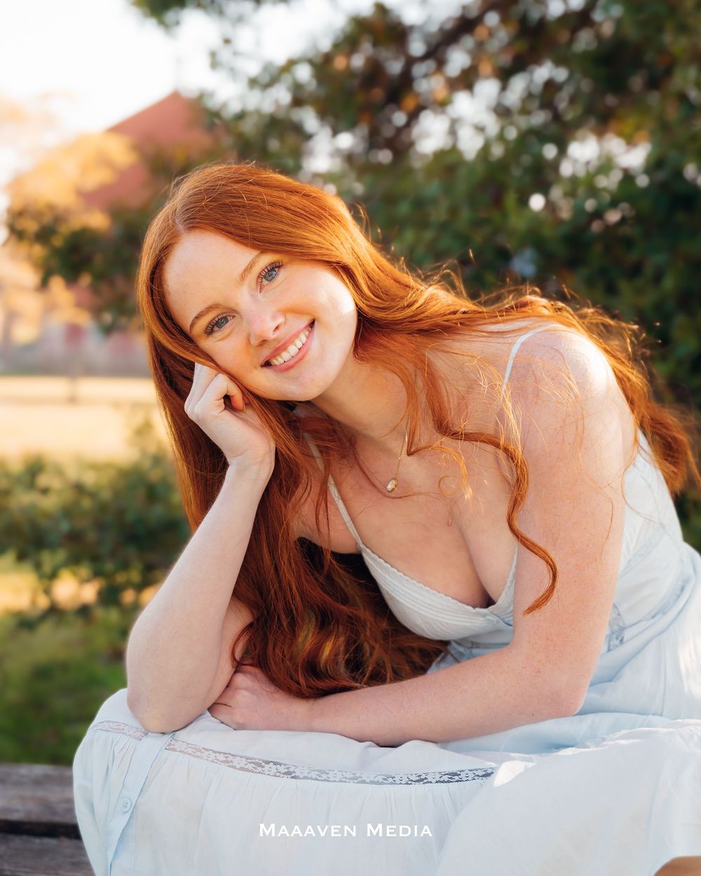 Smiling redhead woman with freckles in a light blue dress, posing outdoors in a sunny park setting.