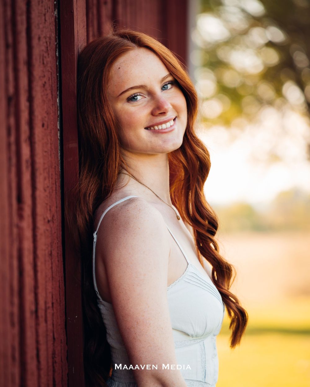 Young woman with long red hair smiling warmly, leaning against a red barn wall in a white dress.