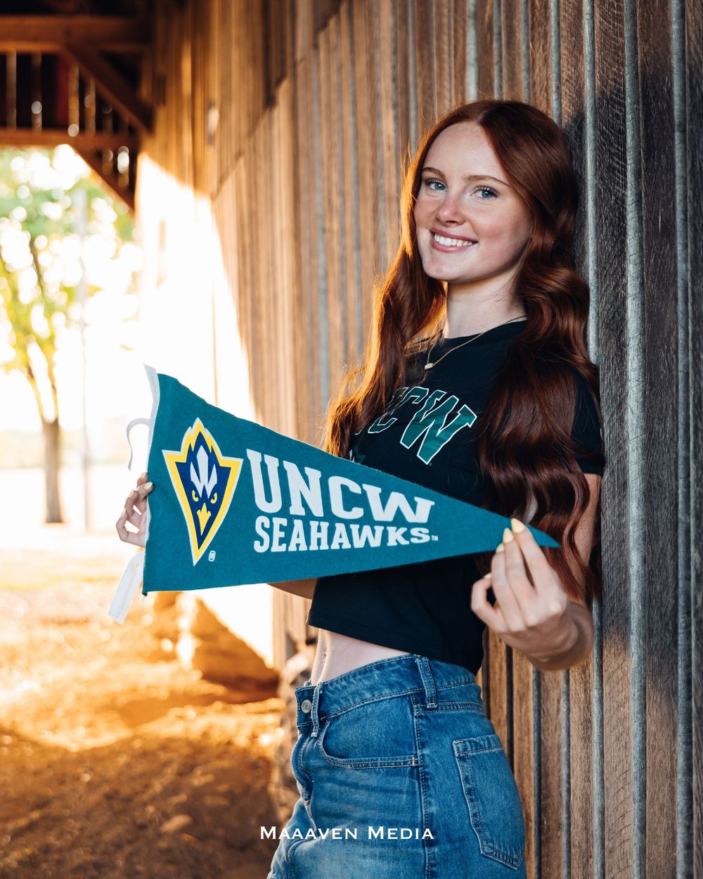 Smiling young woman holding a UNCW Seahawks pennant flag, wearing a black crop top and jeans outdoors.