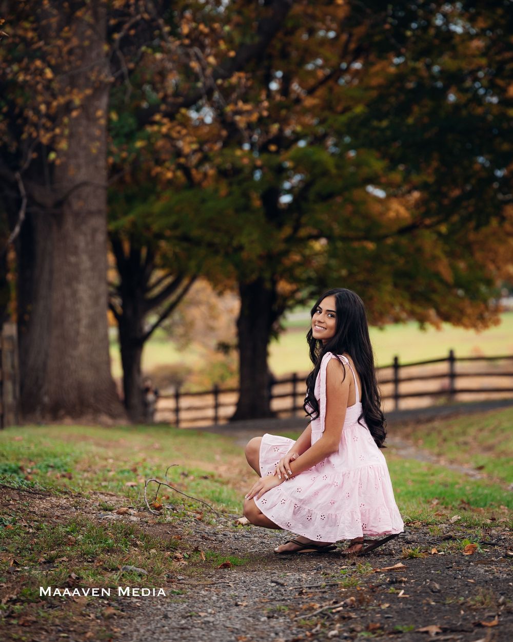 A person in a pink dress sits gracefully on fallen autumn leaves in a park setting with wooden fences and trees.