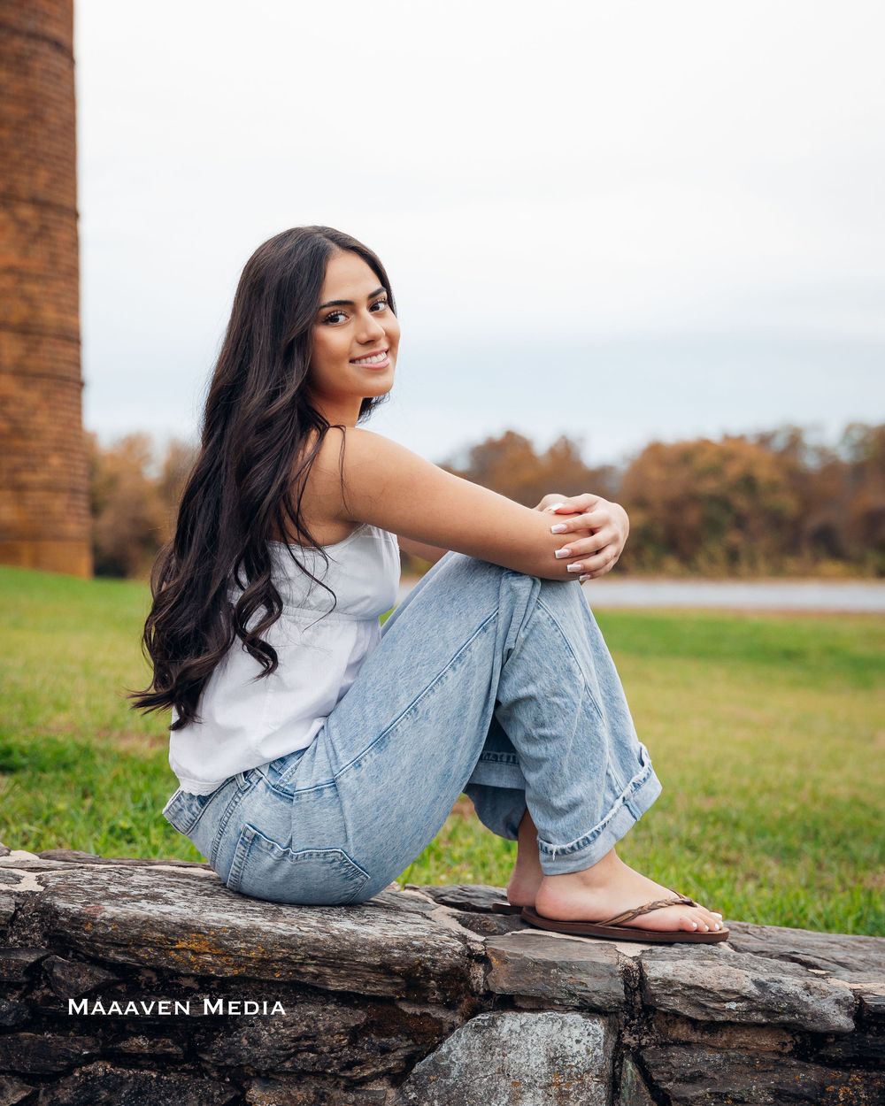 A person in casual attire sits on a fallen log in a grassy field during autumn, with long dark hair and relaxed pose.