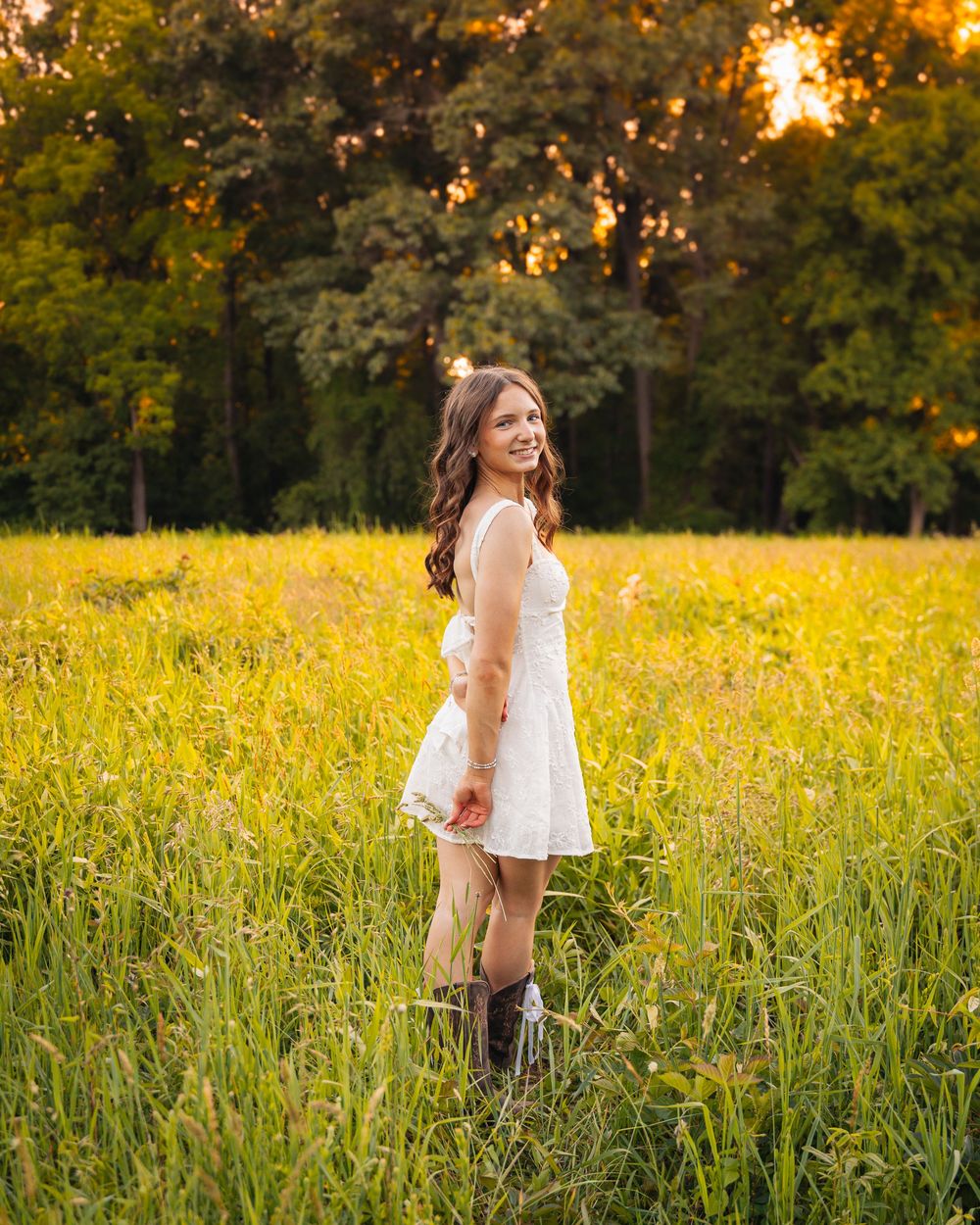 Young woman in white dress and cowboy boots smiling in a sunny green field at golden hour.