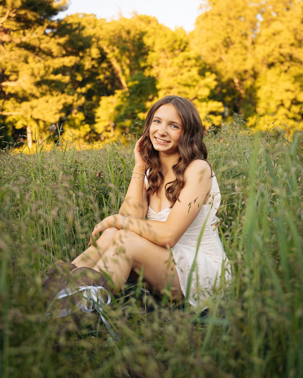 Young woman in white dress sitting in tall green grass field, smiling, with golden sunlit trees in background.