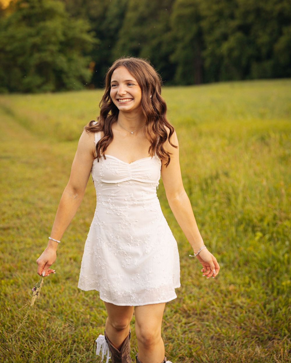 Smiling young woman in white mini dress and cowboy boots walking through a lush green field.