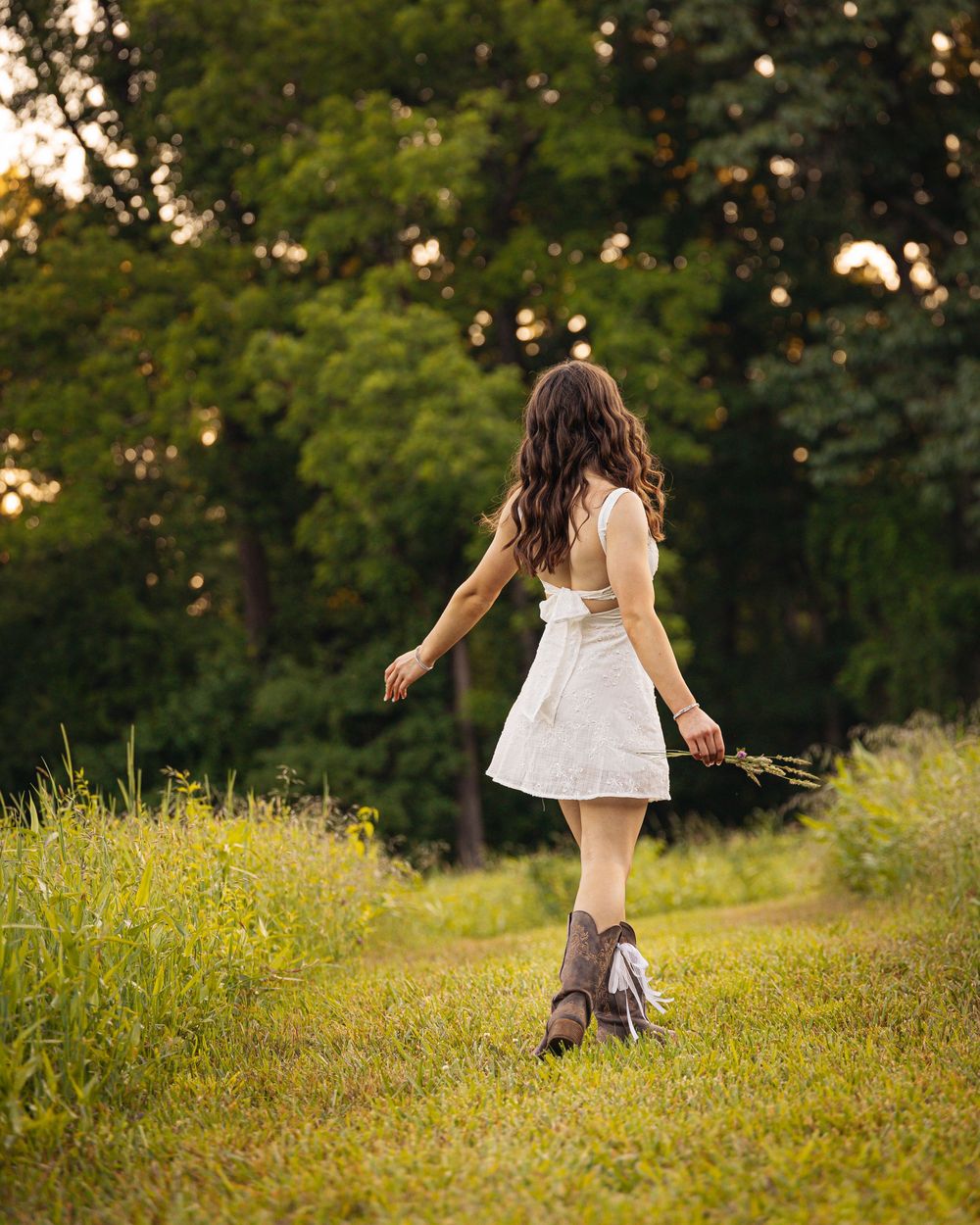 Young woman in white dress and cowboy boots walking through lush green meadow holding wildflowers at golden hour.