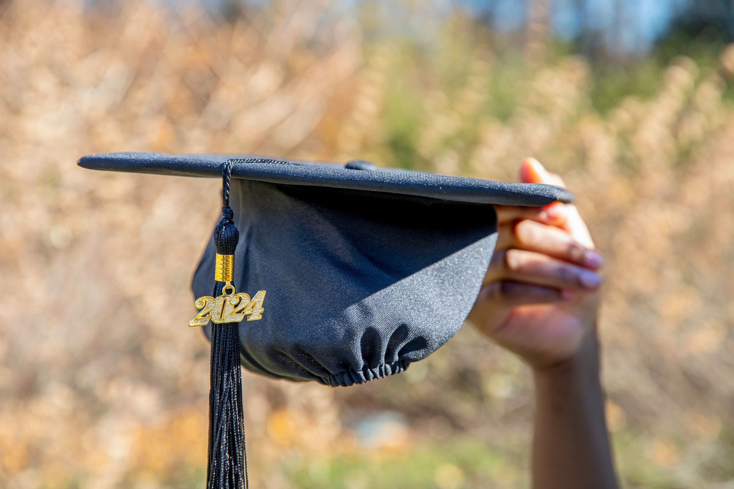 Black graduation cap with gold tassel held up against blurred natural background.