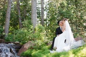 A romantic wedding portrait of newlyweds sitting by a waterfall in a lush forest setting.