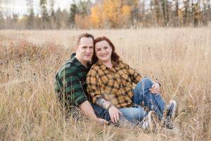 A couple sits together in a golden autumn field surrounded by tall grass and trees.