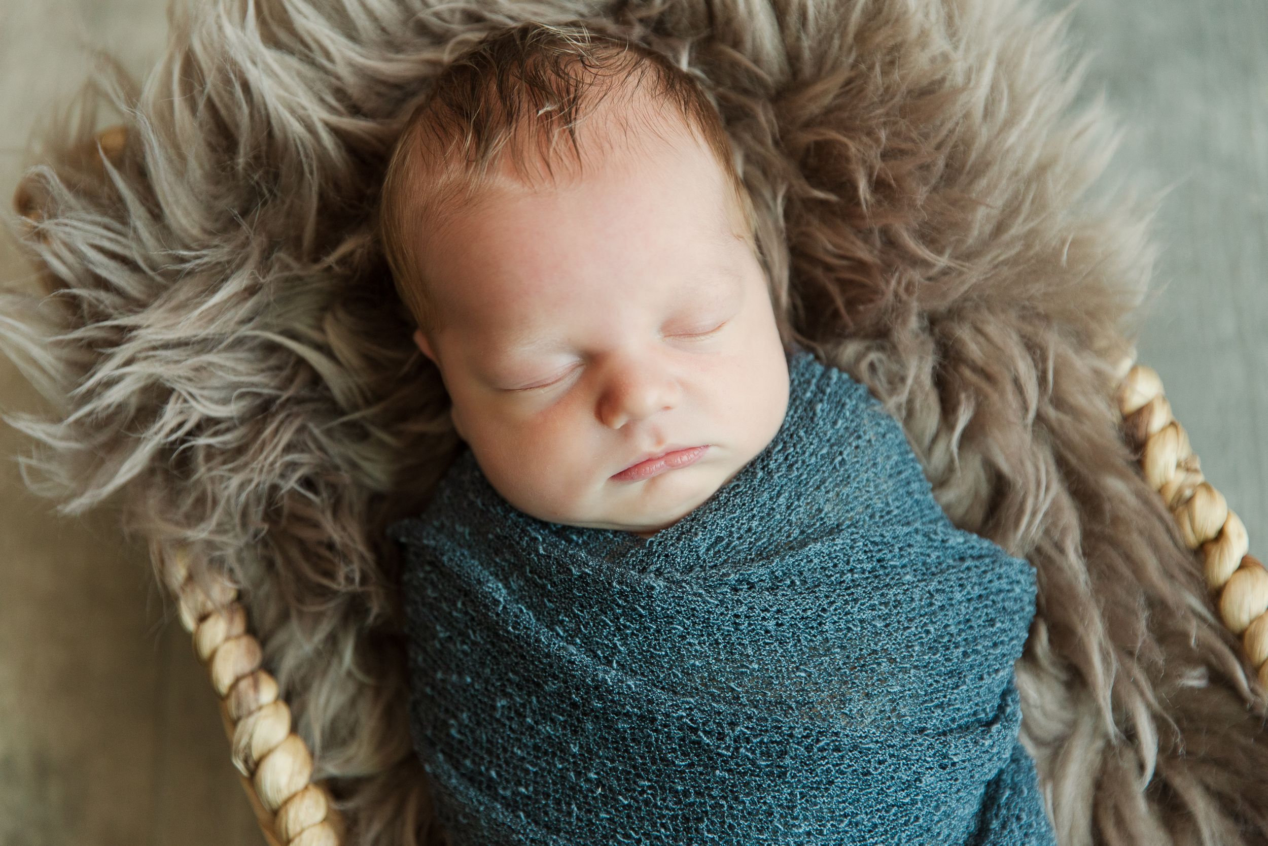 A sleeping newborn wrapped in teal swaddle on fluffy fur in a basket.