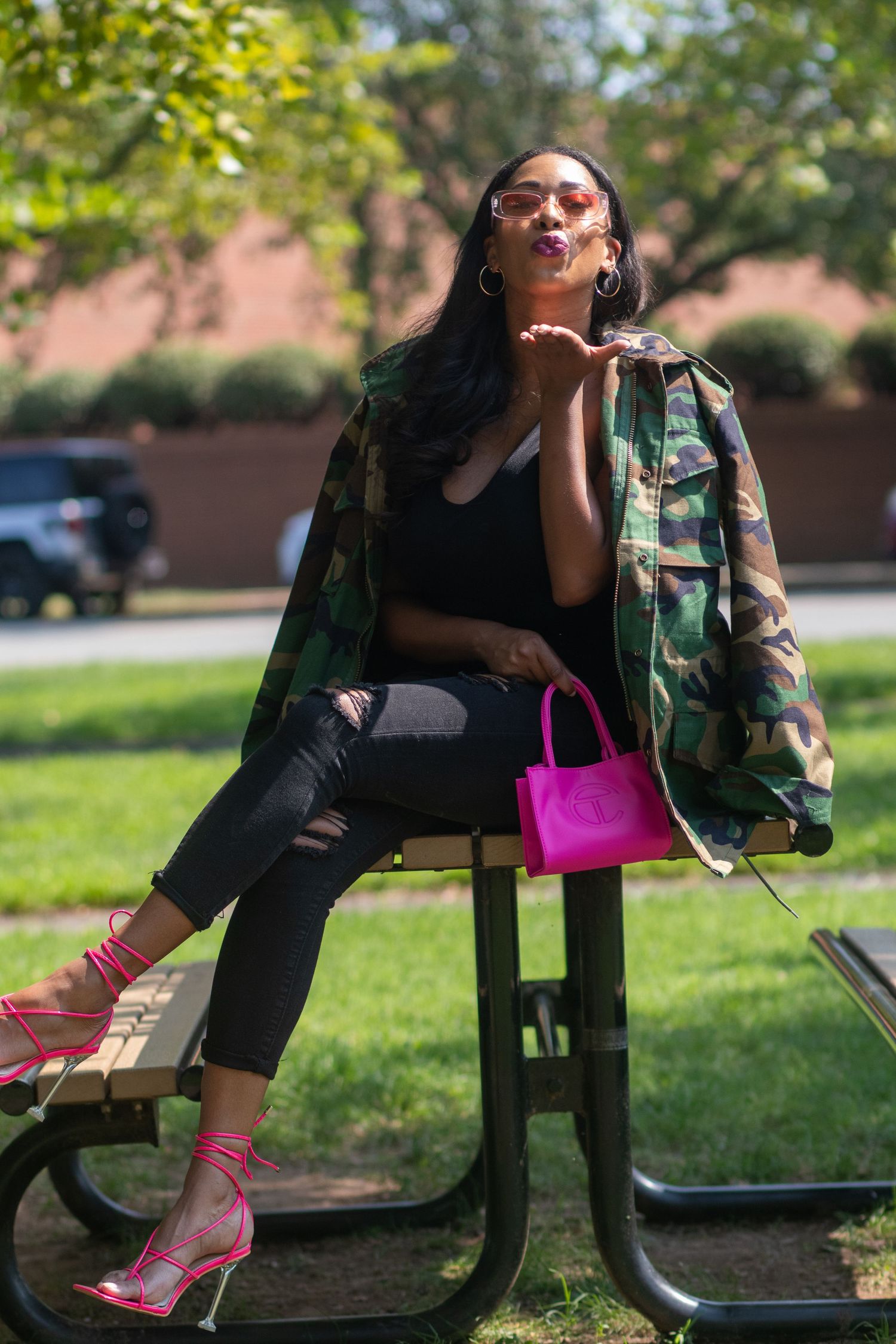 A trendy outfit featuring a camo jacket, black crop top, pink heels and handbag styled casually on a park bench.