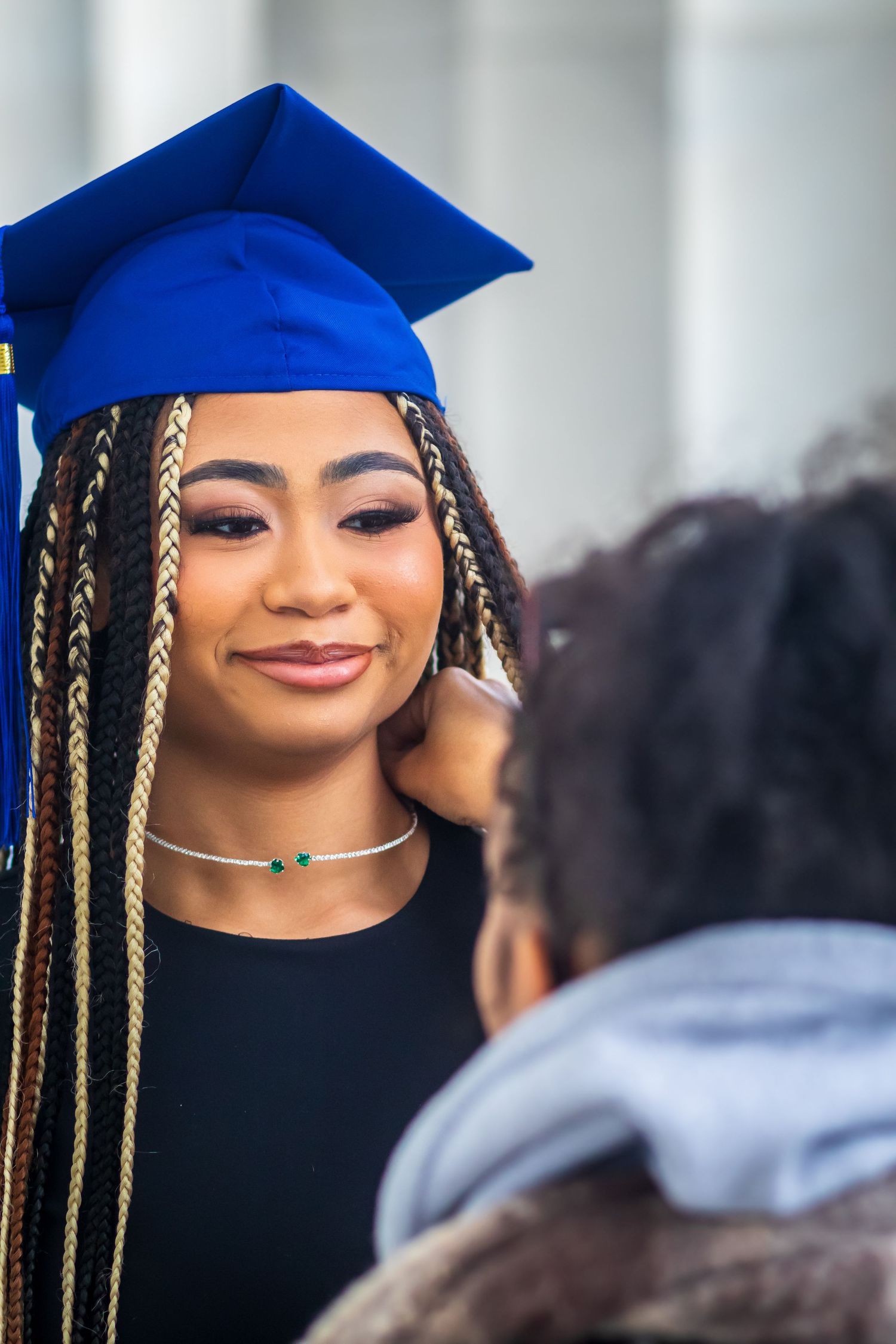 Graduate wearing blue cap with braided hair and delicate necklace during commencement ceremony.