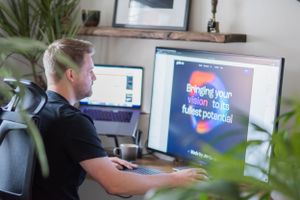 Someone working at a desk with multiple computer monitors in a home office setting with plants visible in the foreground.