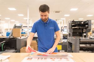 A worker in a blue polo shirt examines printed materials on a workbench in a print production facility.