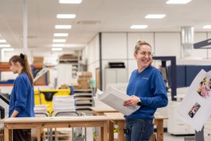 Workers in blue shirts process printed materials at a commercial printing facility with bright fluorescent lighting.