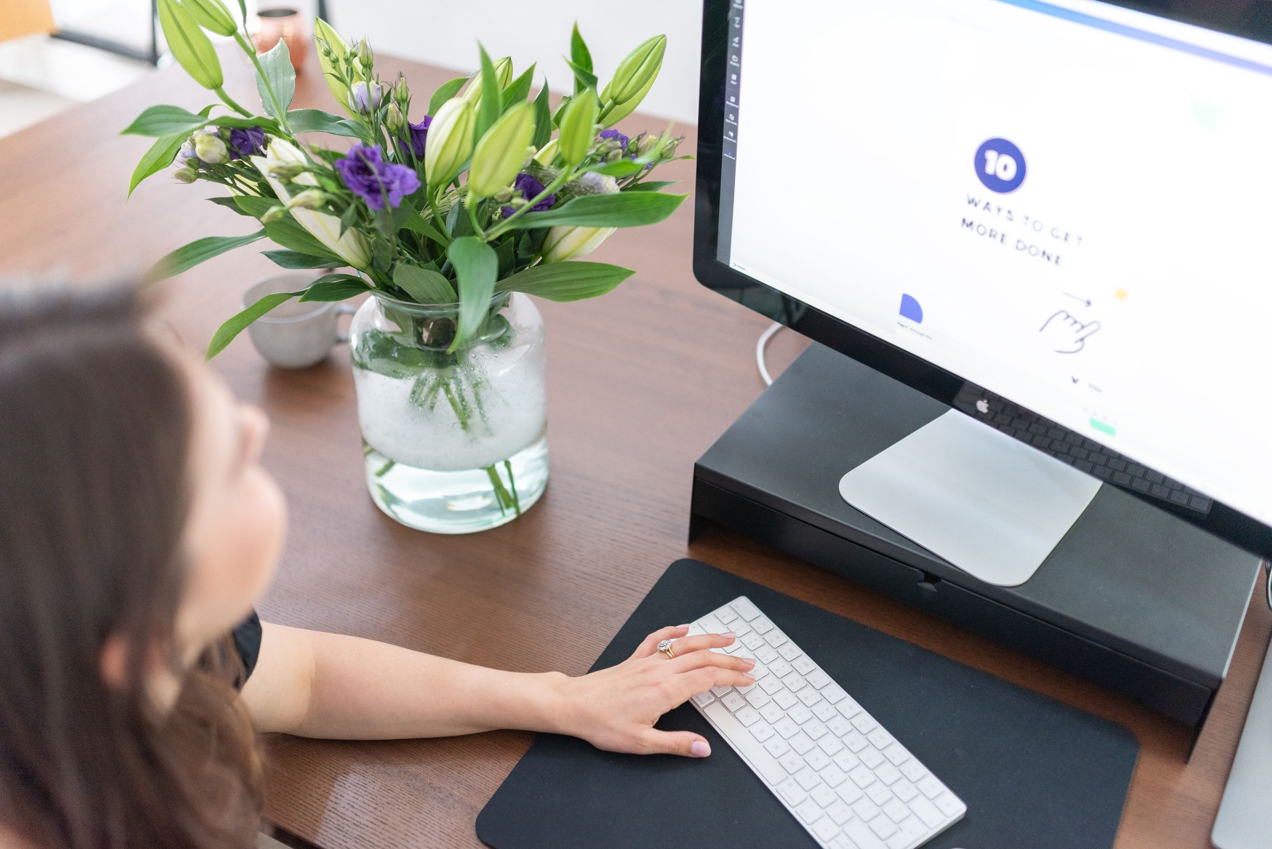 Someone working at a desk with a computer monitor and fresh flowers in a vase nearby.
