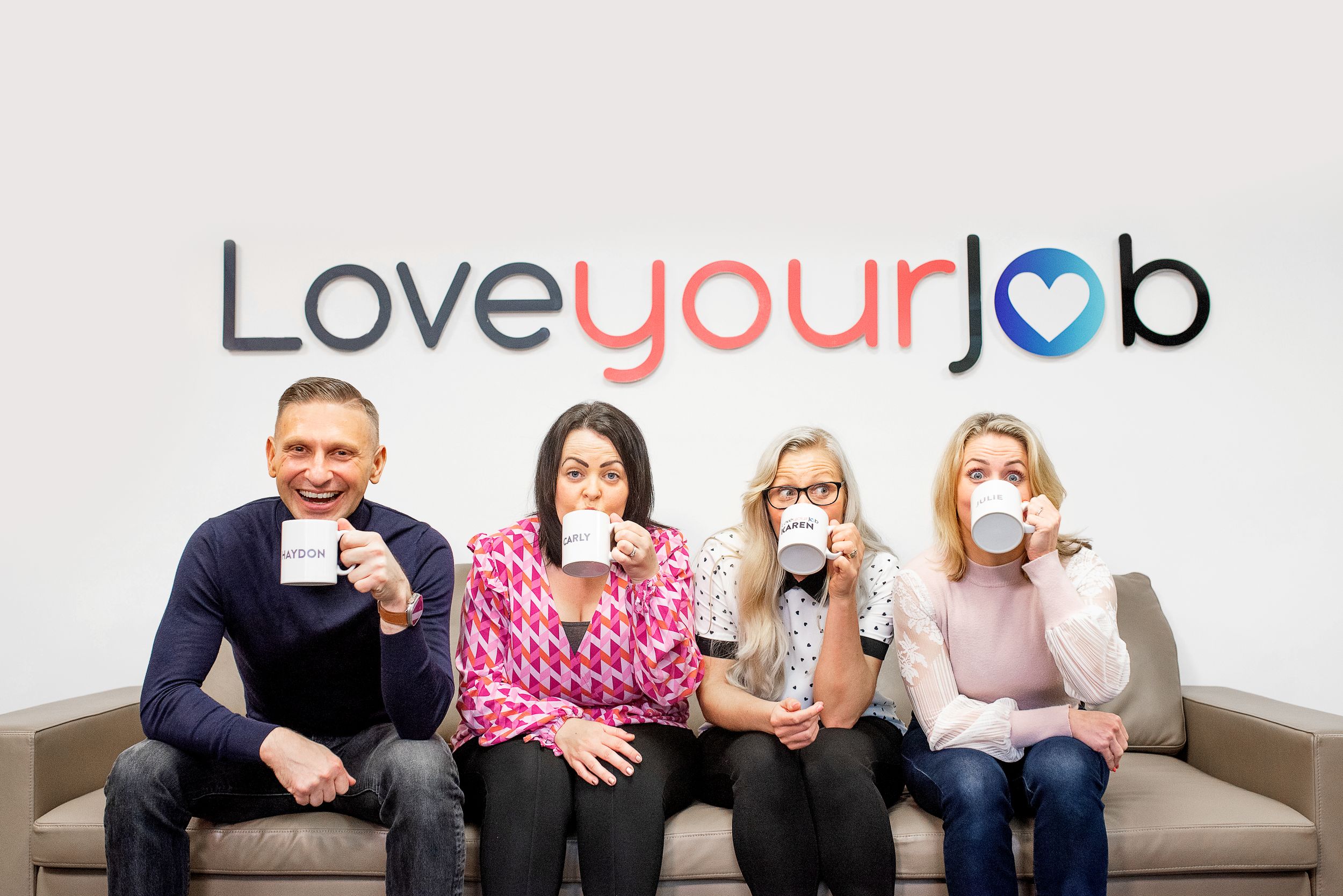 Four coworkers sitting on a couch drinking coffee below a Love Your Job text logo on the wall.