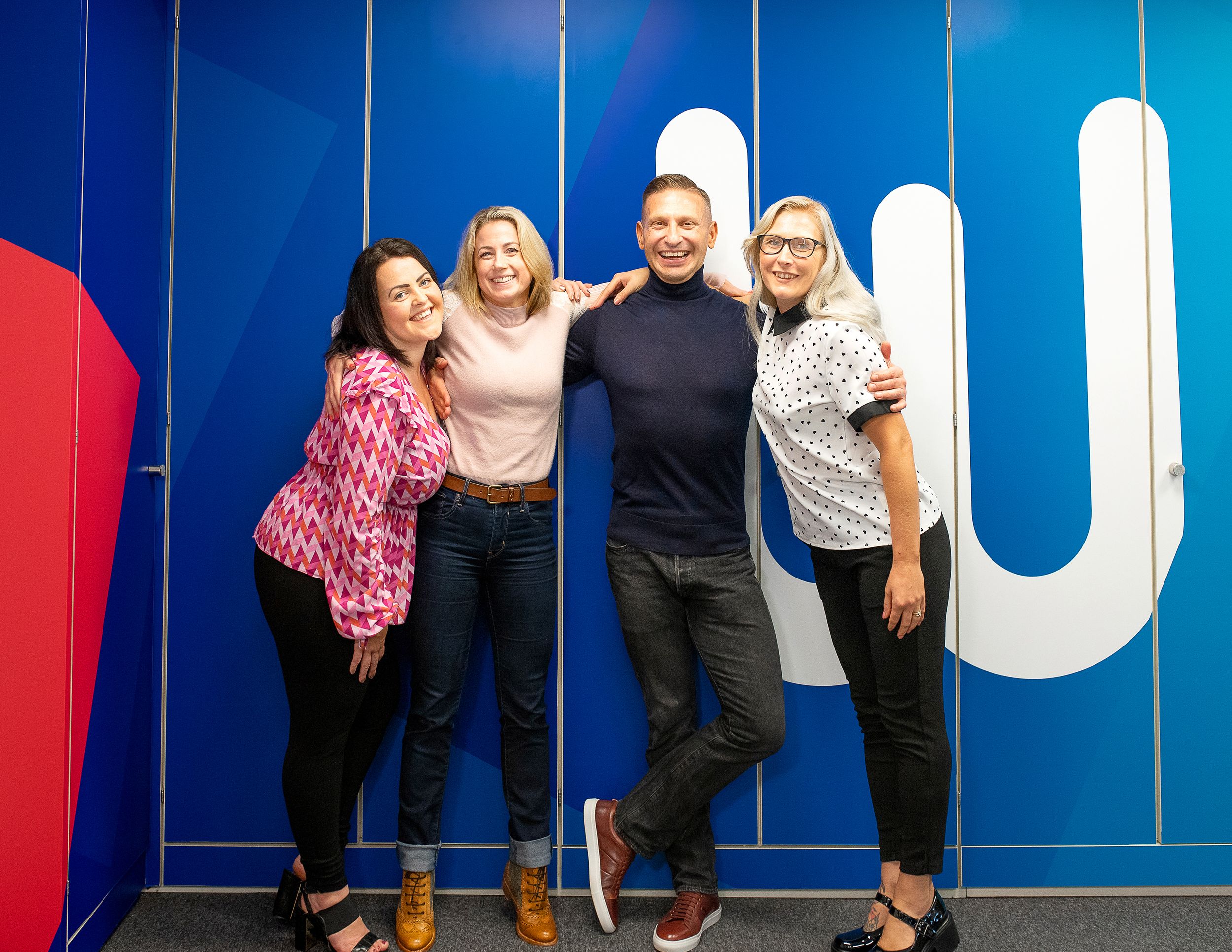 A group poses together against a bright blue wall with white lettering in a modern indoor setting.