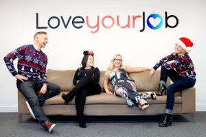 A group of coworkers wearing holiday sweaters relax on a couch under a Love Your Job sign.