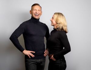 Two people wearing black turtleneck sweaters share a joyful moment against a gray background.