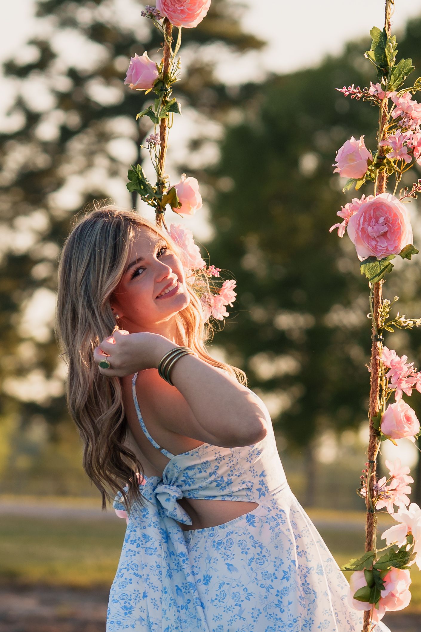 Smiling blonde woman in floral dress on swing decorated with pink roses outdoors at golden hour.