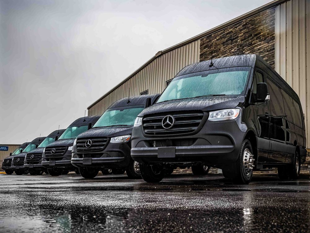A line of black Mercedes-Benz Sprinter vans parked in front of an industrial building on a rainy day.