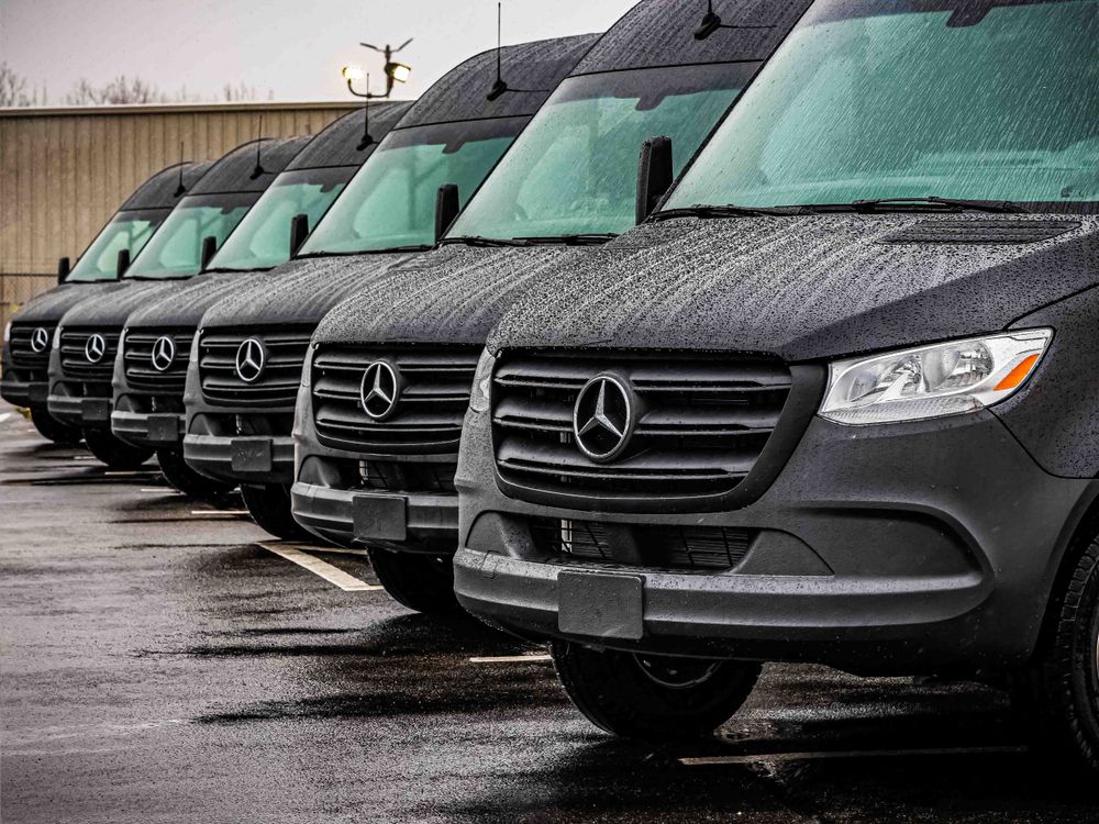 Row of black Mercedes-Benz Sprinter vans lined up in wet parking lot.