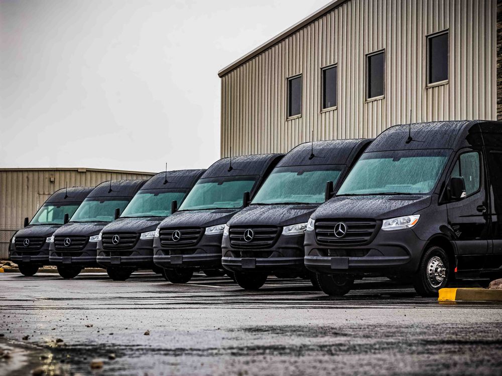 Multiple black Mercedes Sprinter vans lined up in a row against a metal warehouse building.
