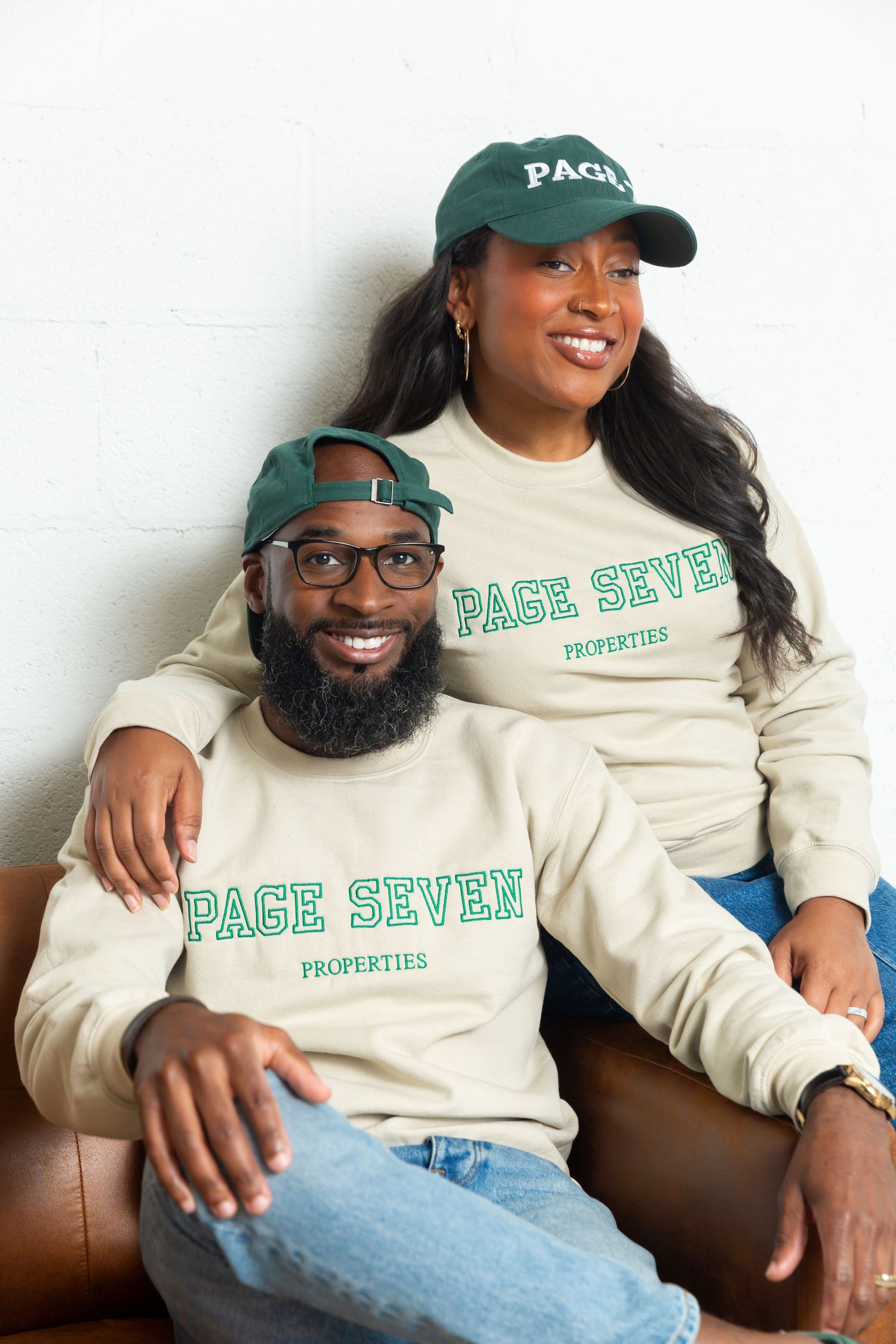 Two people wearing matching cream colored Page Seven sweatshirts and green baseball caps pose together on brown leather seating.
