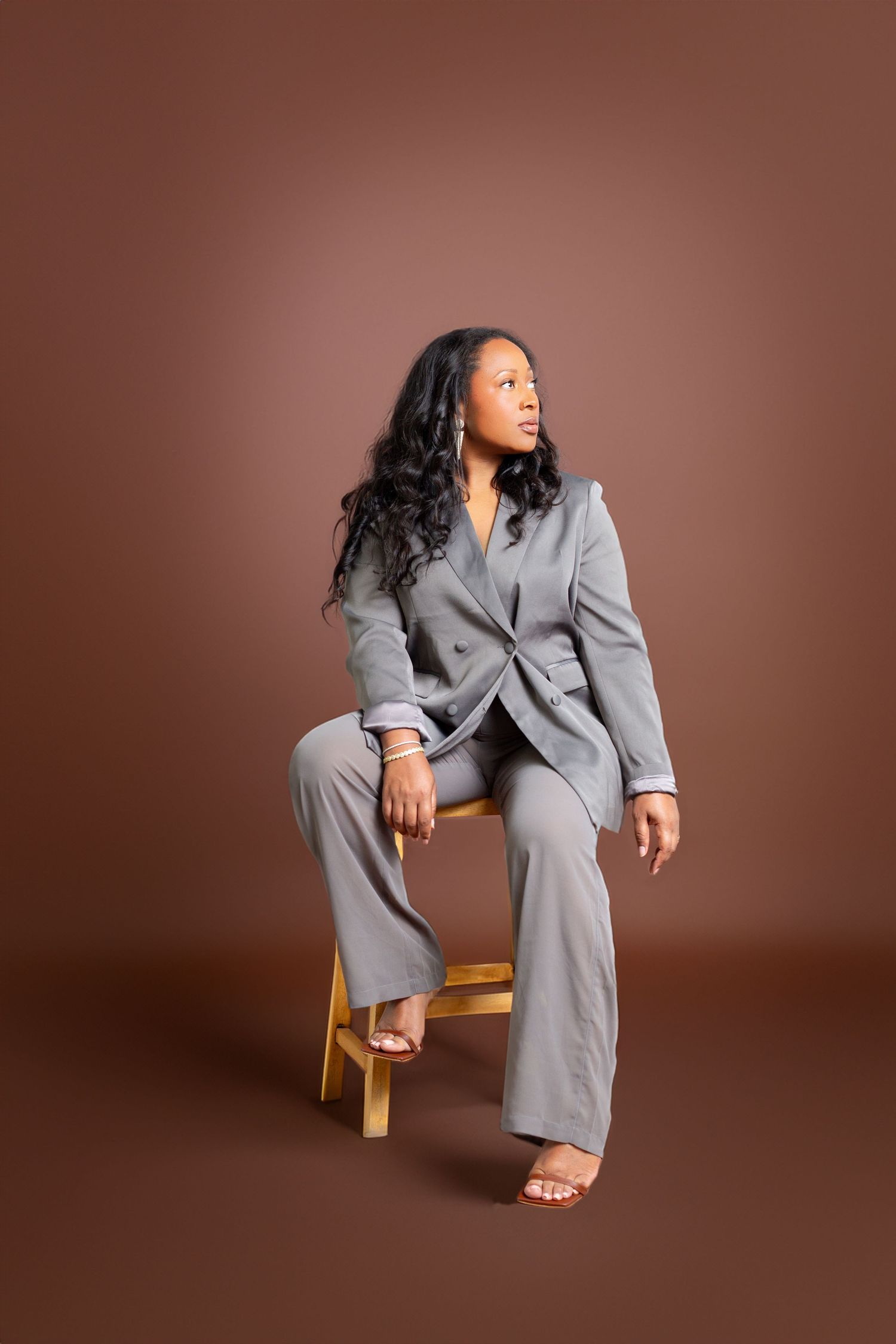Professional portrait of a person in a sleek gray business suit sitting casually on a wooden stool against a brown backdrop.