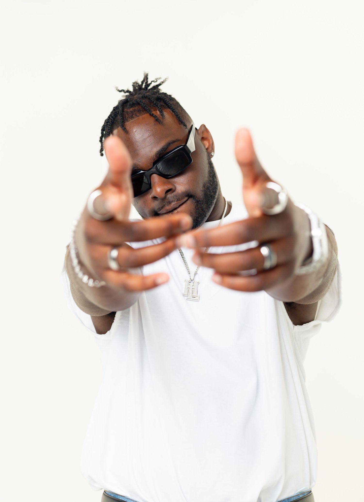 Person in sunglasses wearing a white t-shirt and beige jacket pointing both hands toward the camera against white background.