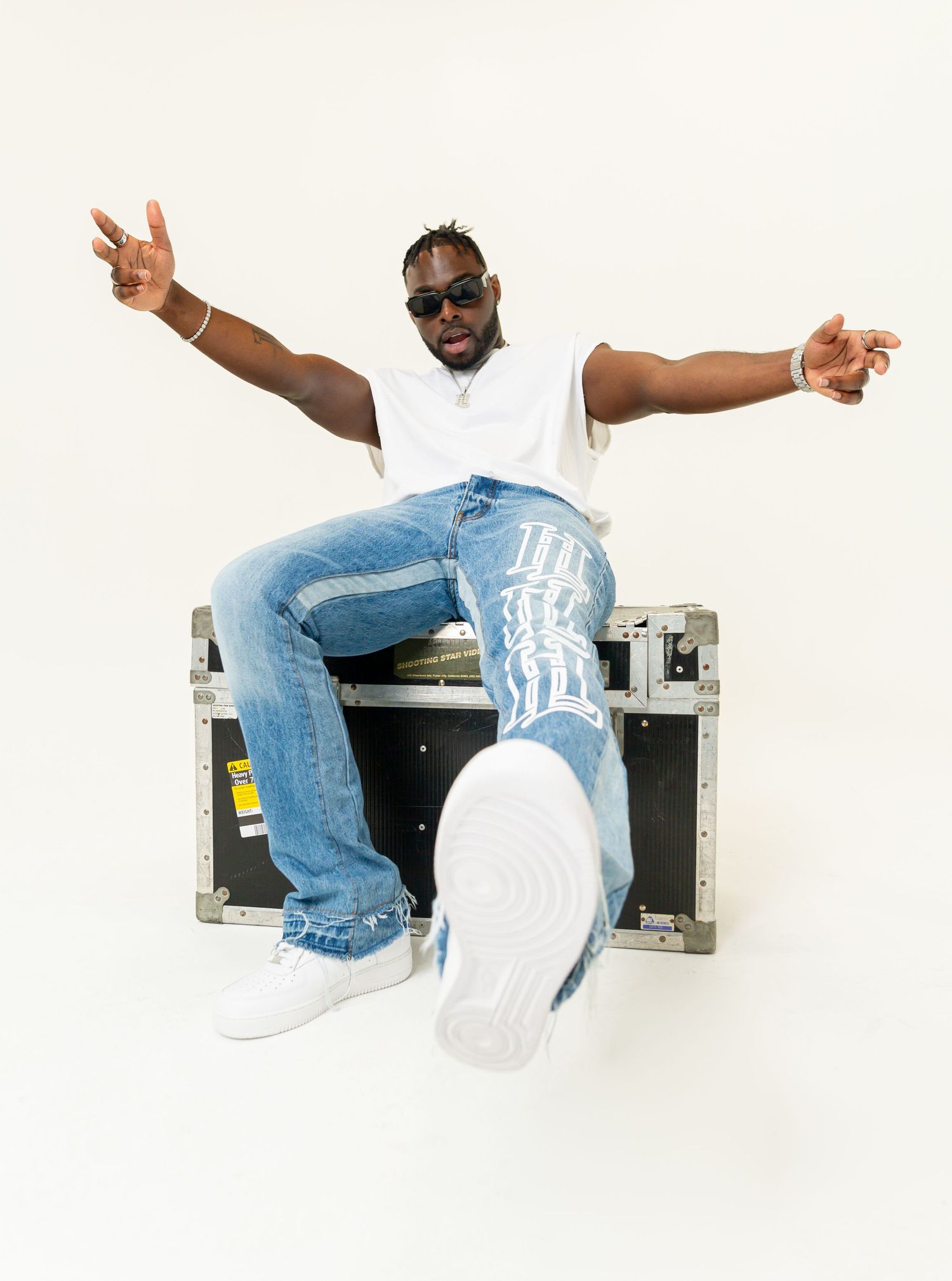 Person in sunglasses and white t-shirt sitting casually on a road case with arms spread wide against white background.