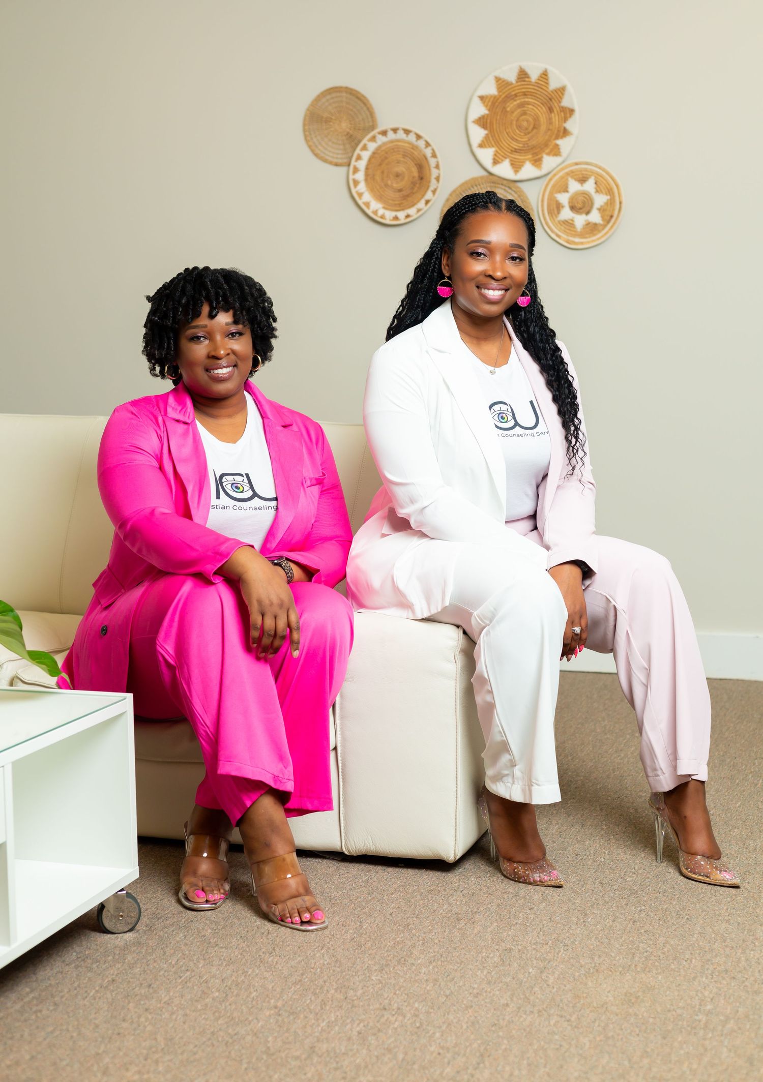 Two women sit together on white furniture wearing matching business attire in pink and white with decorative wall art behind them.