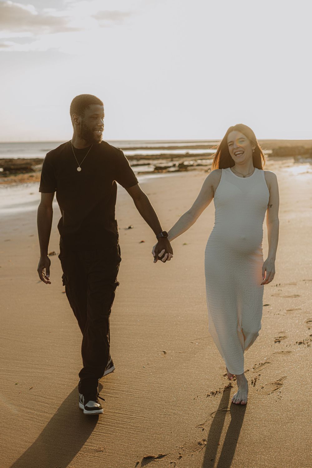 Couple walking hand in hand on beach during golden hour maternity photoshoot.