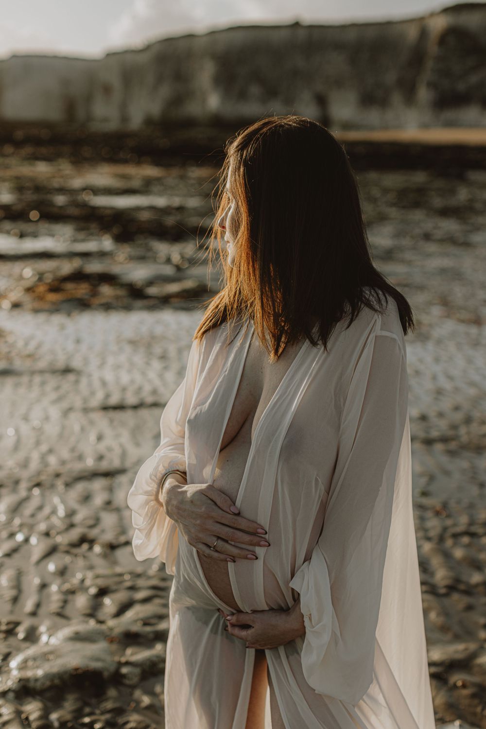 Golden hour maternity photos with flowing white dress on rocky coastline.