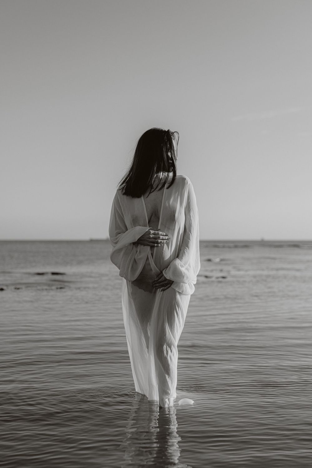 Black and white maternity photography in ocean water with ethereal white dress.