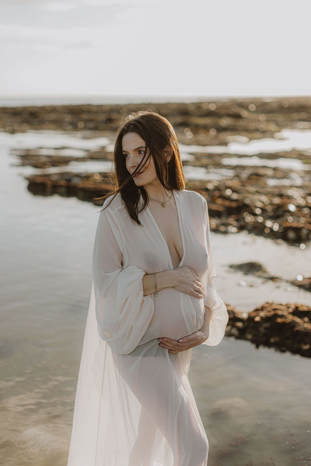 Dreamy coastal maternity photos with flowing white dress at golden hour.