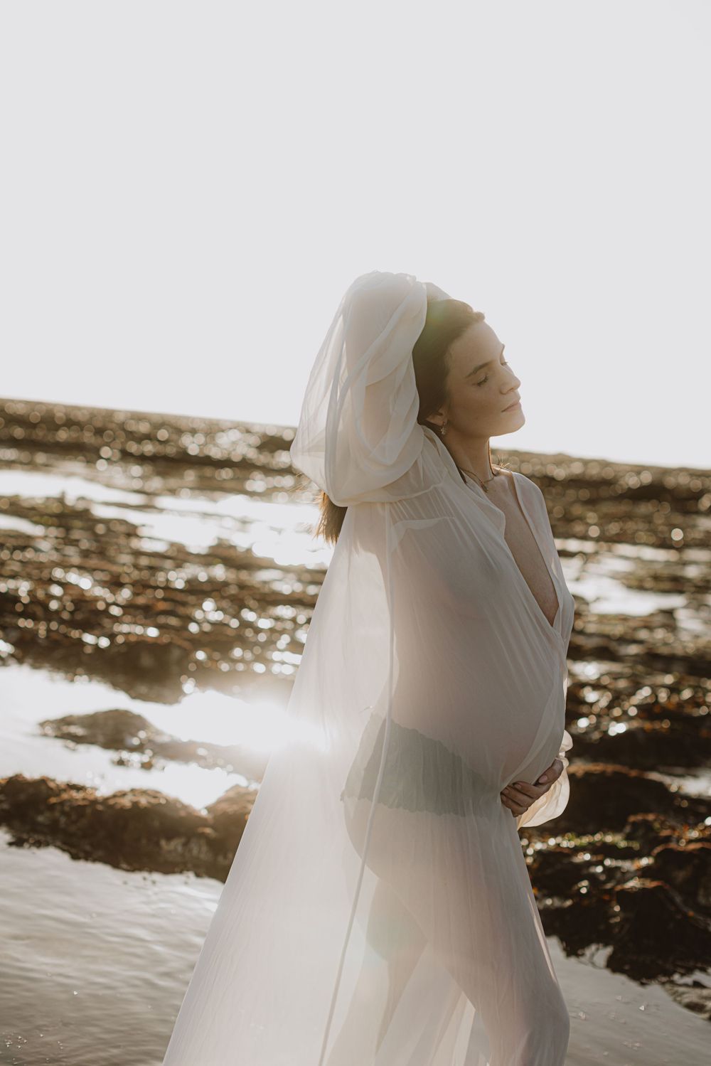 Ethereal beach maternity portraits with backlit white dress at sunset.
