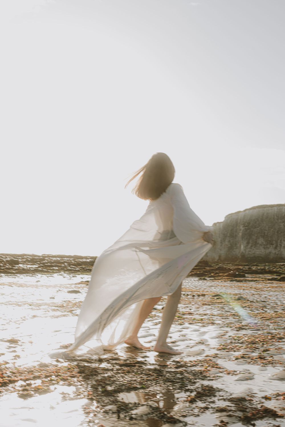 Artistic maternity photos with billowing white dress on rocky shore.