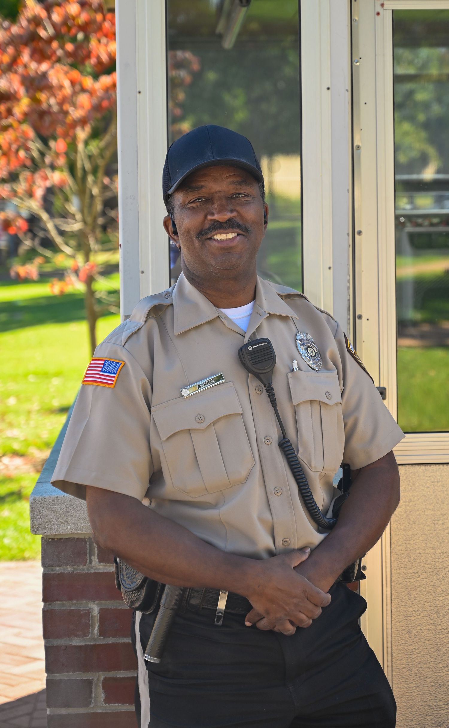 Campus security officer in tan uniform standing at attention by building entrance.