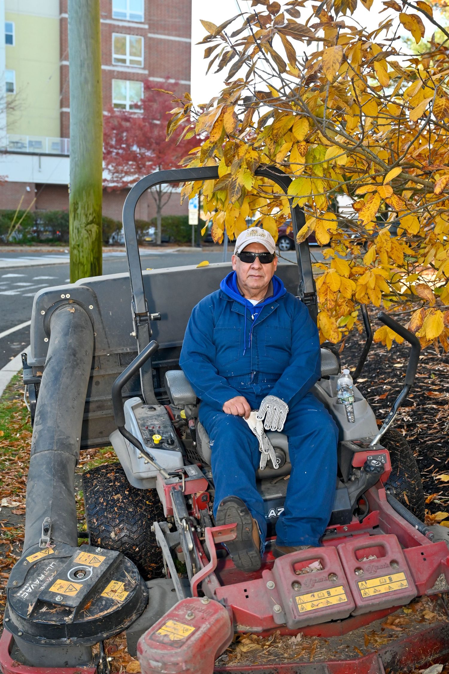 Maintenance worker seated on red commercial lawn mower surrounded by fall foliage.