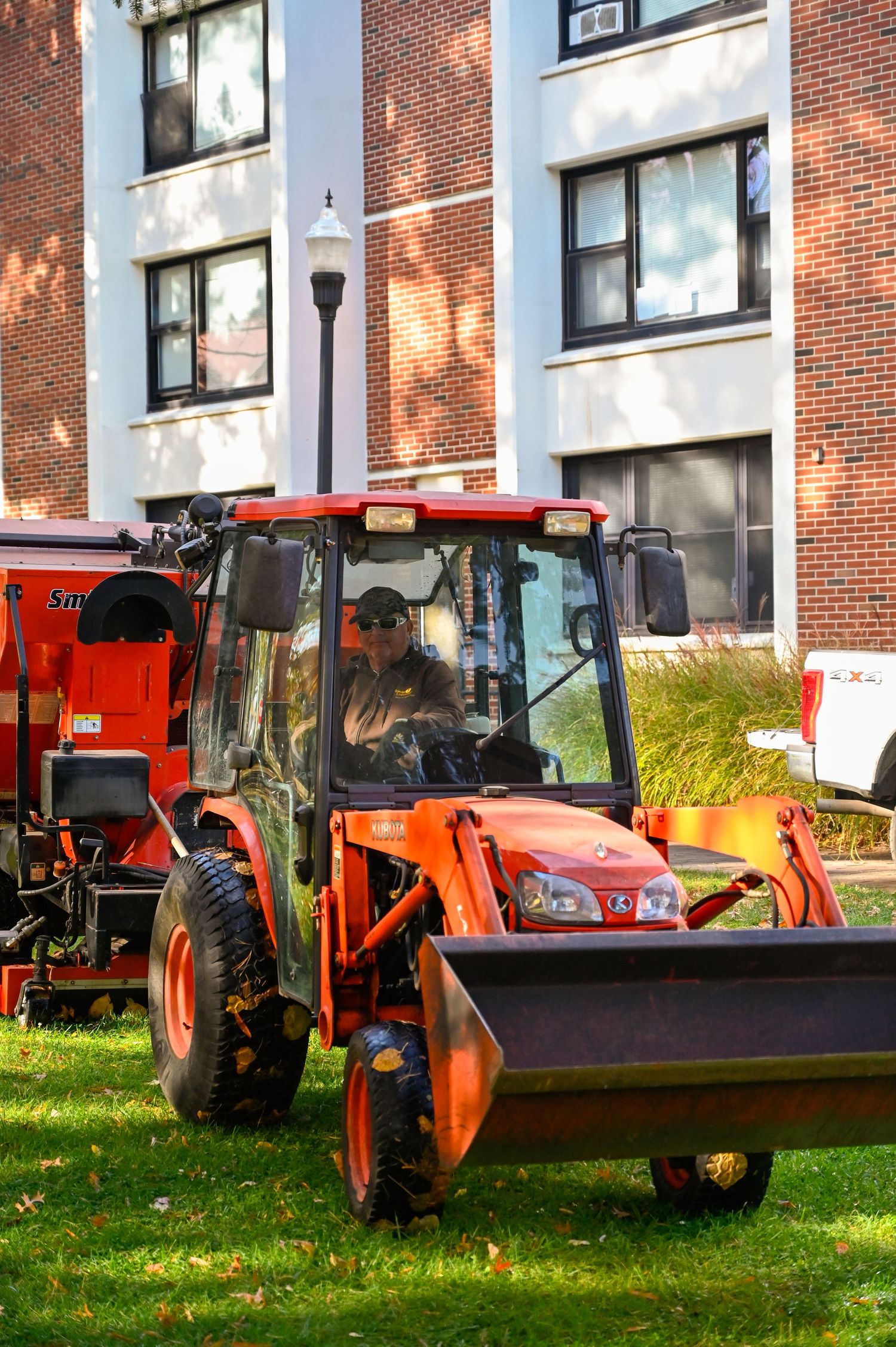 Orange utility tractor with front loader attachment parked on campus lawn near brick building.