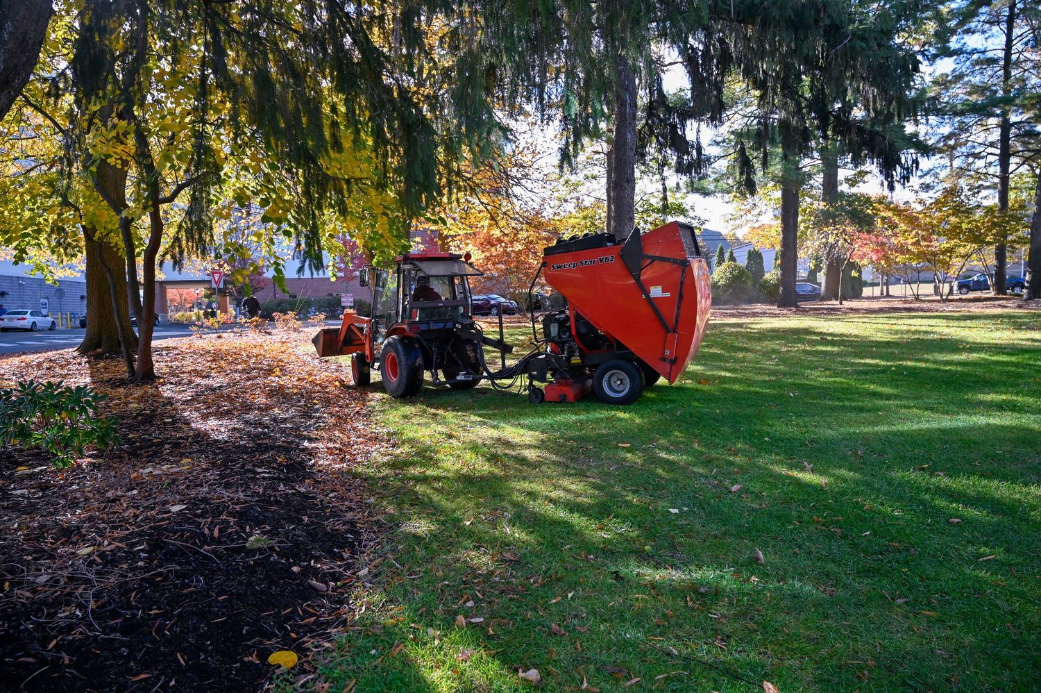 Red maintenance vehicles and equipment working on landscaping beneath autumn trees.