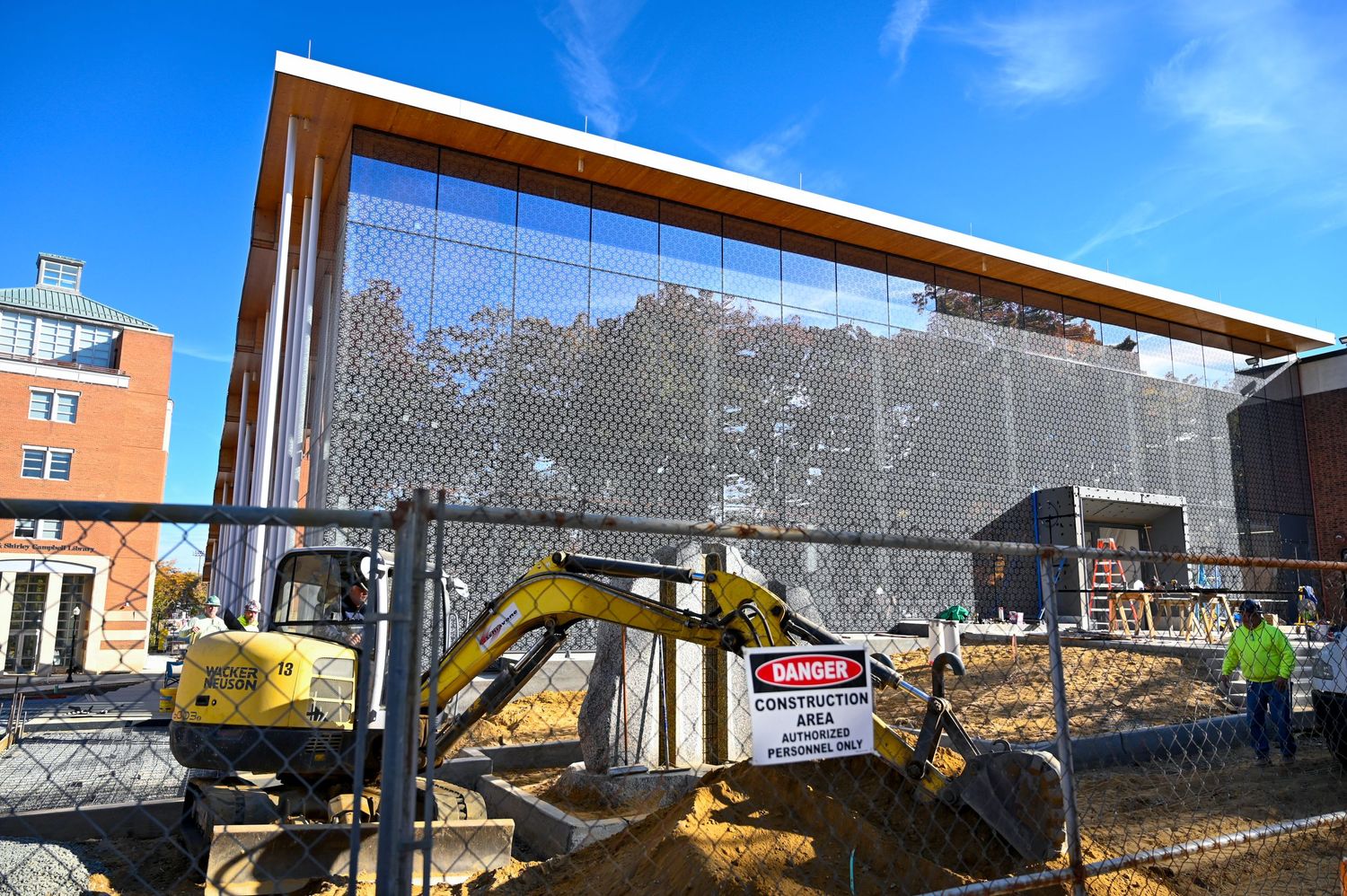 Modern glass building under construction with exposed framework and yellow excavator.