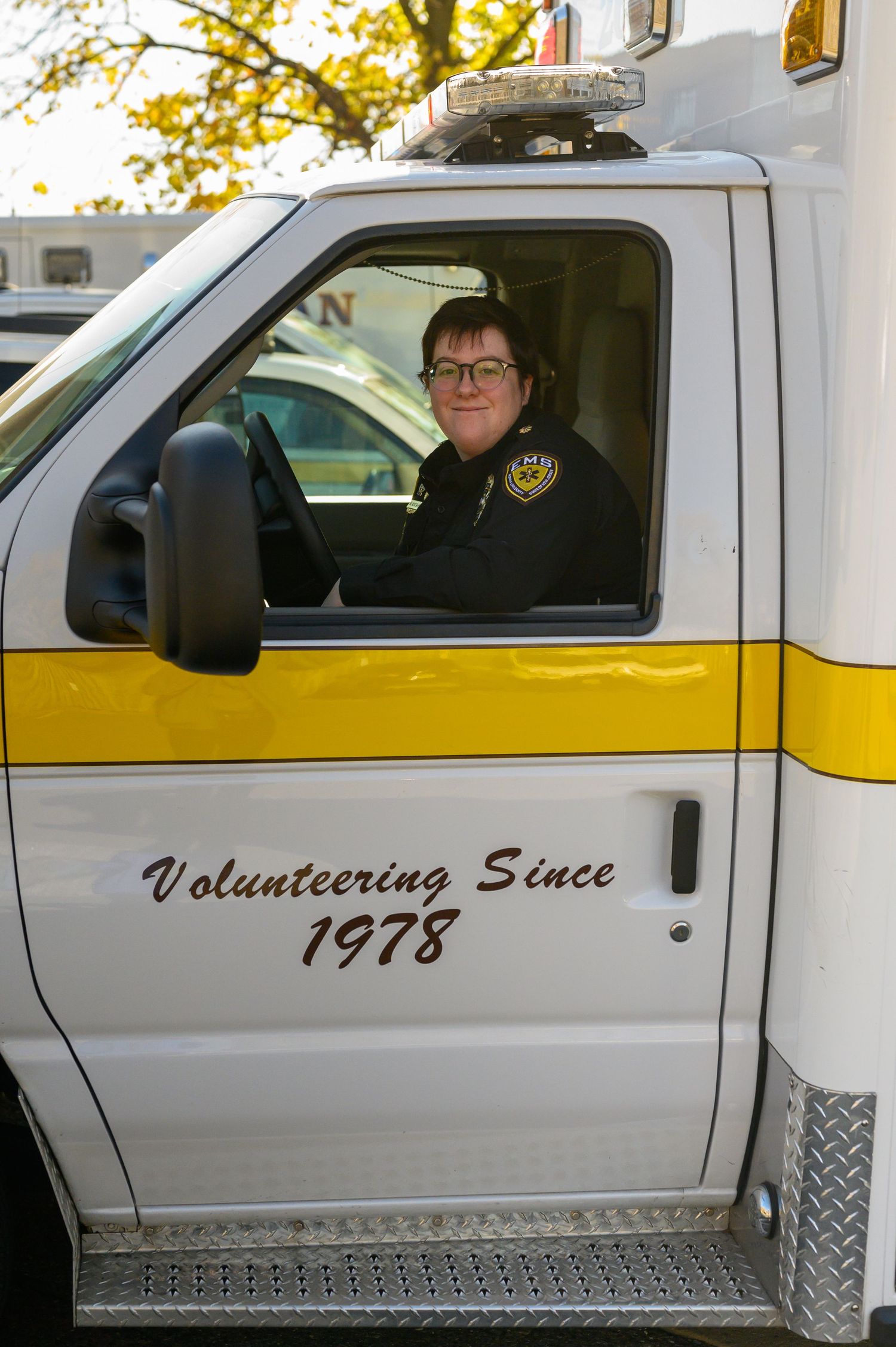 Emergency vehicle with 'Volunteering Since 1978' text on door parked under autumn trees.