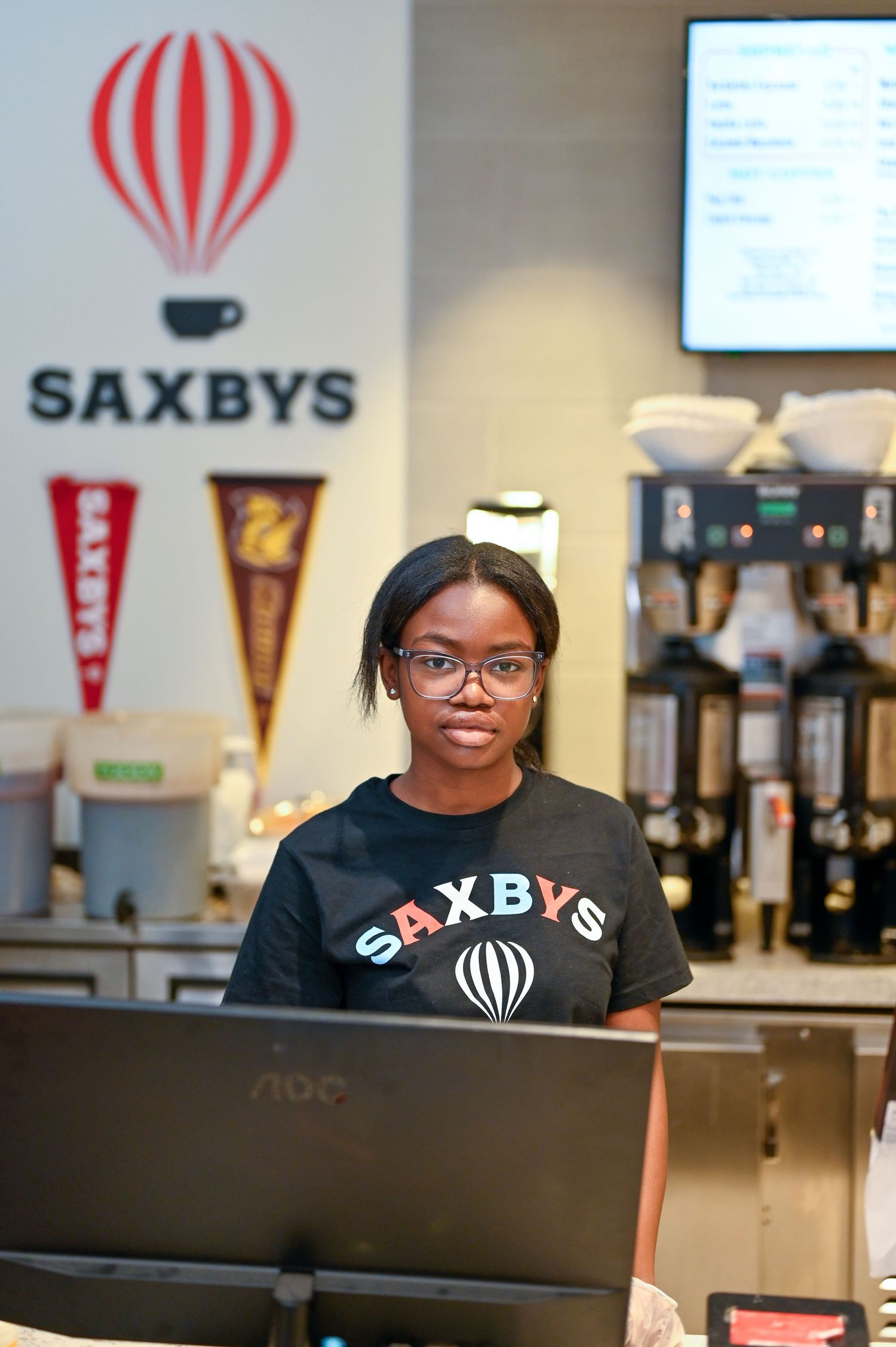 A Saxbys coffee shop employee wearing glasses and a branded black t-shirt stands behind the counter.