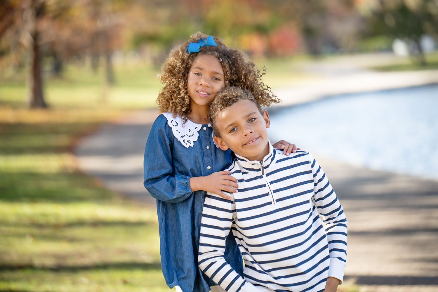 Two siblings pose together on a path in a park, one wearing stripes and one in denim.